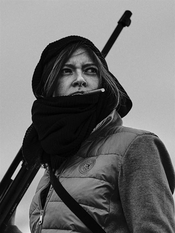 A black and white portrait of a woman in a hood with a rifle over her shoulder