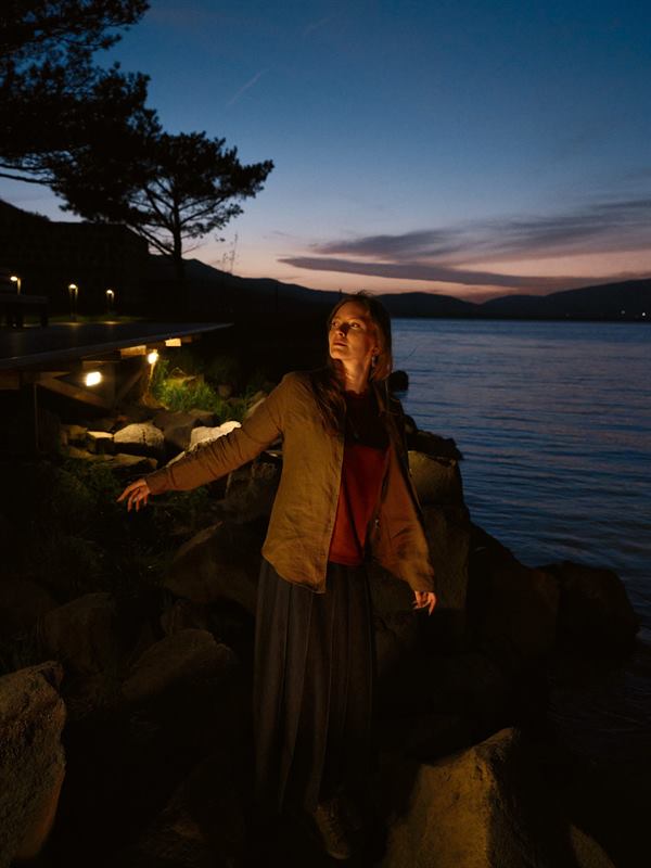 A woman in a yellow coat on rocks by the water at dusk